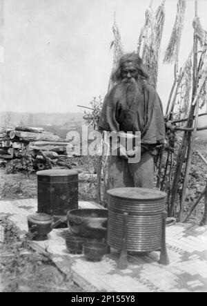 Ainu man standing outdoors on a mat covered with clay containers, 1908. Stock Photo