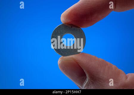 Very small metal washer held between fingers by man. Close up macro studio shot, isolated on blue background. Stock Photo