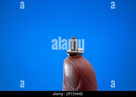 Small metal threaded screw held by Caucasian male hand between fingers. Close up studio macro shot, isolated on blue background. Stock Photo