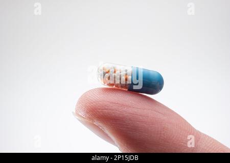 Small medicine medicament capsule half blue half transparent held between fingers. Close up studio macro shot, isolated on white background. Stock Photo
