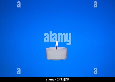 Single small white candle burning. Close up studio shot, isolated on blue background. Stock Photo