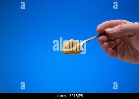 Mustard paste on a small silver spoon held in hand by man. Close up studio shot, isolated on blue background Stock Photo
