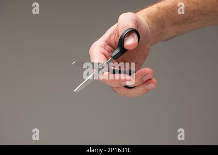 Caucasian male hand holding a small craft scissors with black plastic handles isolated on gray background. Stock Photo