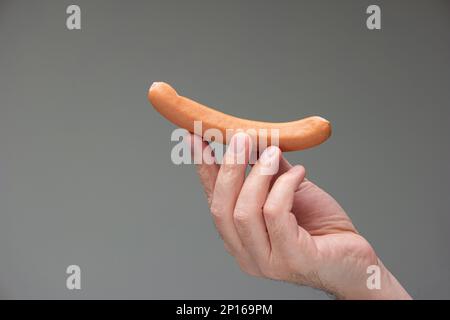 Caucasian male hand holding a bratwurst dog between fingers isolated on gray background. Stock Photo