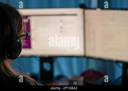 Unrecognizable woman with glasses working from home on two computer screen low key shot from behind. Stock Photo