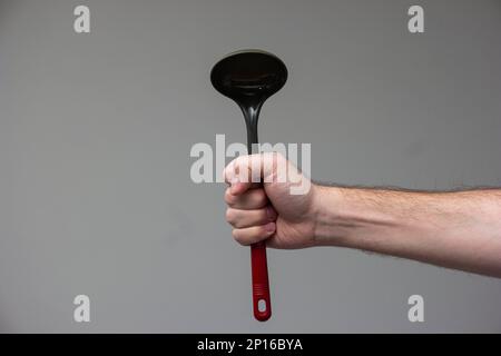 Caucasian male hand holding a black plastic kitchen ladle with red handle isolated on gray background. Stock Photo