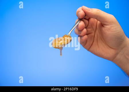 Crystallized honey overflowing from a teaspoon held in hand by Caucasian male hand studio shot isolated on blue background. Stock Photo