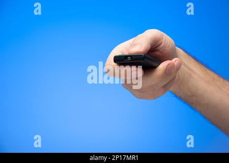 Caucasian male hand holding and pressing buttons on a digital remote control angled towards the camera isolated on blue. Stock Photo