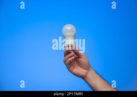 Caucasian male hand holding a white opaque light bulb close up isolated on blue background studio shot. Stock Photo