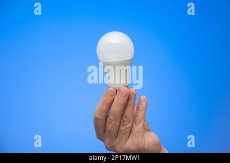 Caucasian male hand holding a white opaque light bulb close up isolated on blue background studio shot. Stock Photo