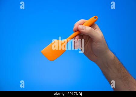 Caucasian male hand holding orange silicon spatula isolated on blue background. Stock Photo