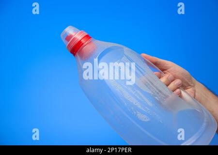 Caucasian male hand holding a large empty plastic bottle with dew and droplets upside down isolated on blue background studio shot. Stock Photo