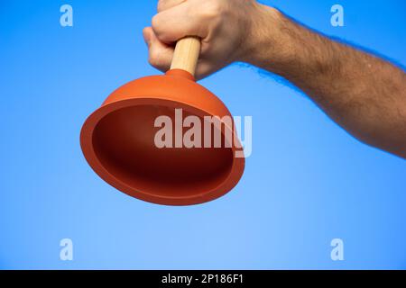 Orange rubber bathroom plunger held in hand by Caucasian male hand close up studio shot isolated on blue. Stock Photo