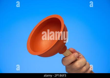 Orange rubber bathroom plunger held in hand by Caucasian male hand close up studio shot isolated on blue. Stock Photo