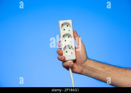 Electrical extension cord with multiple plug sockets held in hand by Caucasian male hand isolated on blue. Stock Photo