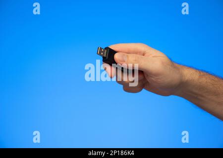 Small USB memory stick plug held in hand by Caucasian male hand isolated on blue. Stock Photo