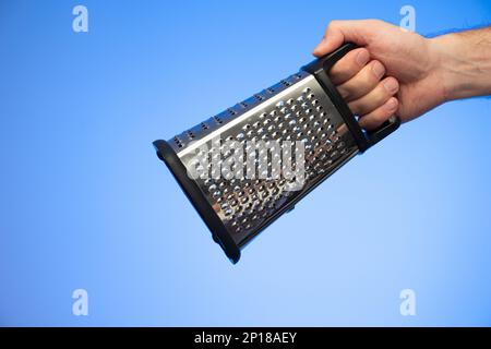 Stainless metal kitchen grater with plastic handle held in hand by Caucasian male hand studio shot isolated on blue background. Stock Photo