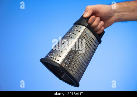 Stainless metal kitchen grater with plastic handle held in hand by Caucasian male hand studio shot isolated on blue background. Stock Photo