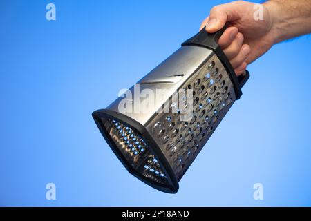 Stainless metal kitchen grater with plastic handle held in hand by Caucasian male hand studio shot isolated on blue background. Stock Photo