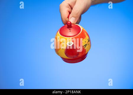 Painted decorative ceramic tea pot held in hand by Caucasian male hand studio shot isolated on blue background. Stock Photo