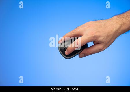 Small black plastic wireless computer mouse held in hand by Caucasian male hand studio shot isolated on blue background. Stock Photo
