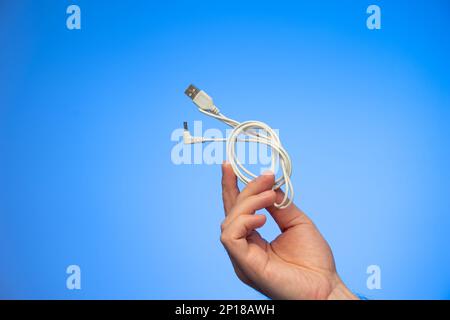 White folded USB charging cable held in hand by Caucasian male hand studio shot isolated on blue background. Stock Photo