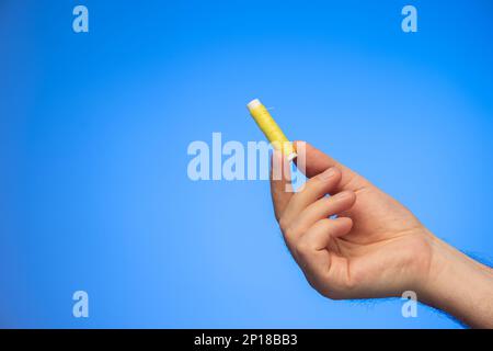 Small reel of yellow sewing thread held in hand by Caucasian male hand studio shot isolated on blue background. Stock Photo