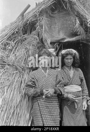 Two Ainu women standing at the entrance of a hut, 1908. Stock Photo