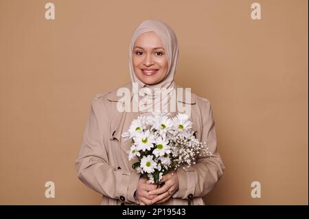 Beautiful middle-aged Muslim woman in elegant beige coat and hijab, posing with chamomilles on isolated cream background Stock Photo