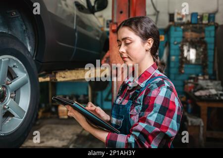 Portrait of a young female mechanic in uniform using a tablet. In the background is an auto repair shop. Stock Photo