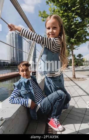 well dressed girl in denim vest and jeans standing near metallic fence next to boy on riverside,stock image Stock Photo