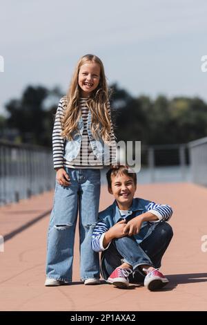 full length of smiling girl in denim outfit standing near happy preteen boy on river embankment,stock image Stock Photo