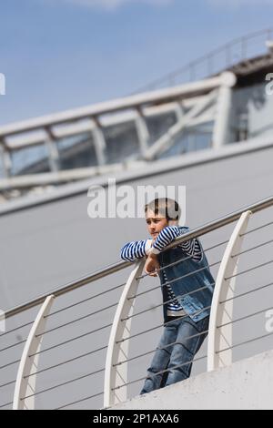 low angle view of preteen boy in denim vest and jeans leaning on metallic fence on embankment,stock image Stock Photo