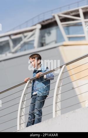 low angle view of stylish boy in denim vest and jeans standing near metallic fence on embankment,stock image Stock Photo