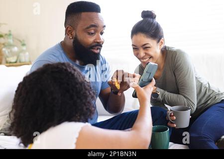 Happy diverse friends sitting on sofa, drinking coffee and using smartphone Stock Photo