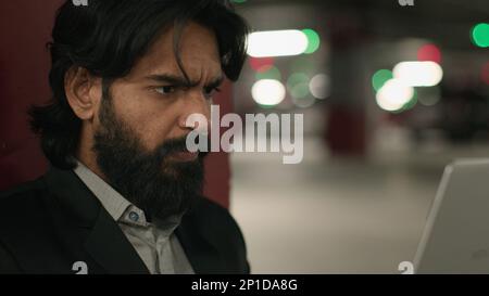 Close-up puzzled serious Indian man looking at laptop screen in underground parking focused worried young arabian businessman feeling anxiety from Stock Photo