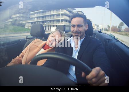 Middle aged couple riding convertible car on urban road. People driving carefree in landscape city  Stock Photo