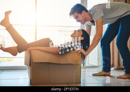 They dont take themselves too seriously. Shot of a young man pushing his girlfriend around in a box while they move into their new home together. Stock Photo