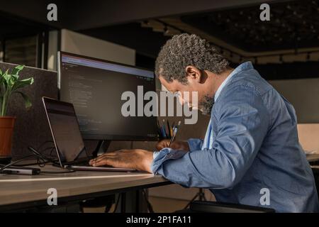 Black Man Writing Lines of Code On Desktop PC With Two Monitors and a Laptop Aside in Stylish Office. Professional Male Developer Programming Stock Photo