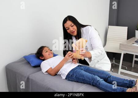 Latino pediatrician doctor checks her girl patient with autism spectrum disorder ASD, she communicates through his teddy bear that she carries as an a Stock Photo