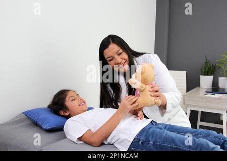 Latino pediatrician doctor checks her girl patient with autism spectrum disorder ASD, she communicates through his teddy bear that she carries as an a Stock Photo