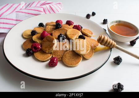 The appearance of tiny pancakes with honey, raspberries and blackberries on a white plate. Stock Photo