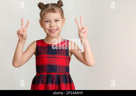 Smiling child little girl showing sign V Victory on both hands caucasian kid of 6 7 years in red plaid dress on grey background Stock Photo