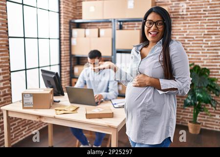 Young hispanic woman expecting a baby working at small business ecommerce celebrating achievement with happy smile and winner expression with raised h Stock Photo