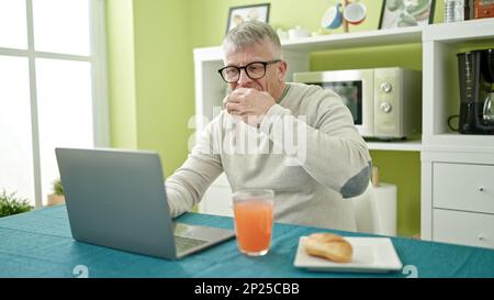 Middle age grey-haired man having breakfast using laptop at home Stock Photo