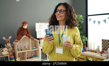 Middle age hispanic woman preschool teacher using smartphone drinking coffee at kindergarten Stock Photo
