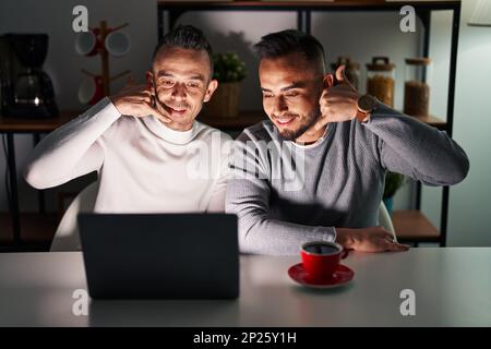 Homosexual couple using computer laptop smiling doing phone gesture with hand and fingers like talking on the telephone. communicating concepts. Stock Photo