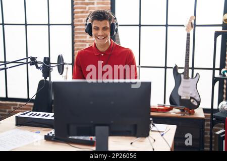 Young hispanic man playing piano keyboard at music studio looking positive and happy standing and smiling with a confident smile showing teeth Stock Photo
