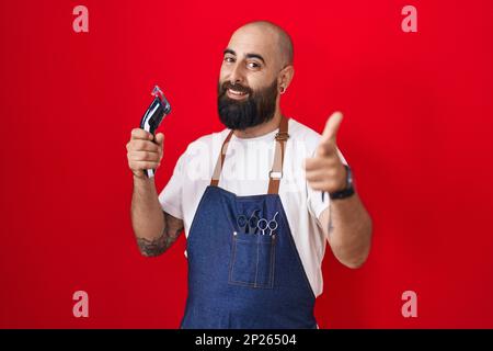 Young hispanic man with beard and tattoos wearing barber apron holding razor pointing fingers to camera with happy and funny face. good energy and vib Stock Photo