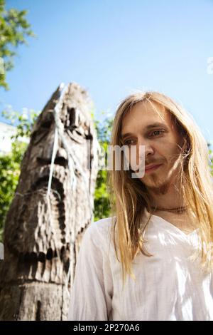 Slavic pagan young long-haired man next to a wooden pole idol sculpture. Traditional Russian culture - religion and art Stock Photo